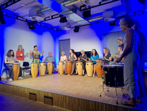 Eine Gruppe von Menschen, meist Frauen, sitzt und steht im Halbkreis auf einer Bühne und spielt Conga-Trommeln und eine kleine Trommel unter blauem Licht in einem modernen Raum mit Holzfußboden und einer industriellen Decke.