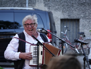 Anreas Koll mit Brille und Weste lächelt, während er im Freien ein Akkordeon spielt und in ein Mikrofon singt. Im Hintergrund sind ein schwarzer Lieferwagen und ein Fahrrad zu sehen.