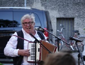 Anreas Koll mit Brille und Weste lächelt, während er im Freien ein Akkordeon spielt und in ein Mikrofon singt. Im Hintergrund sind ein schwarzer Lieferwagen und ein Fahrrad zu sehen.