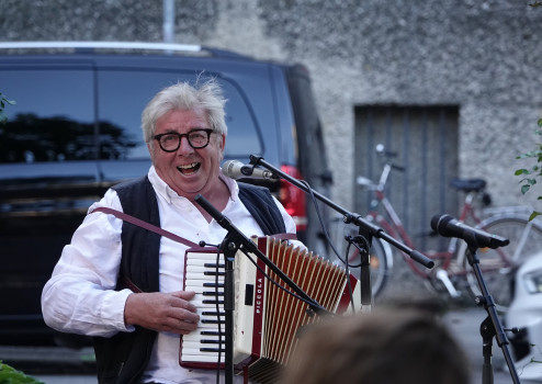 Anreas Koll mit Brille und Weste lächelt, während er im Freien ein Akkordeon spielt und in ein Mikrofon singt. Im Hintergrund sind ein schwarzer Lieferwagen und ein Fahrrad zu sehen.