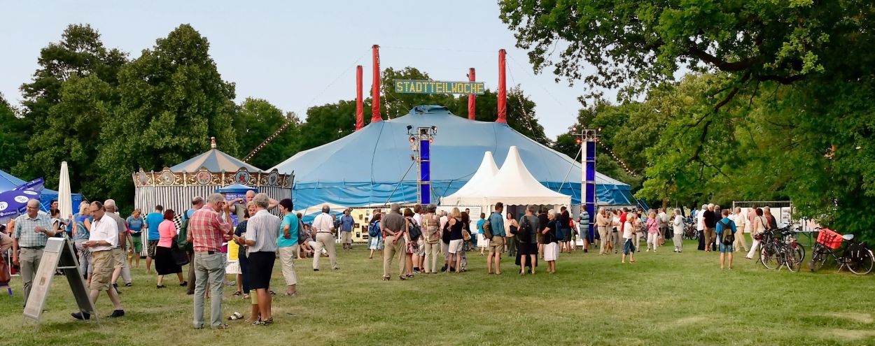 Eine Menschenmenge versammelt sich auf dem Festplatz vor Zirkuszelt, Karussell und Pavillons, mit Bäumen im Hintergrund und geparkten Fahrrädern in der Nähe.