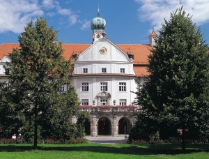 Ein historisches weißes Gebäude mit rotem Ziegeldach, Uhr und grüner Kuppel, teilweise von Bäumen verdeckt und umgeben von einem üppigen grünen Rasen unter einem blauen Himmel mit Wolken.