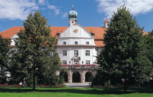 Ein historisches weißes Gebäude mit rotem Ziegeldach, Uhr und grüner Kuppel, teilweise von Bäumen verdeckt und umgeben von einem üppigen grünen Rasen unter einem blauen Himmel mit Wolken.