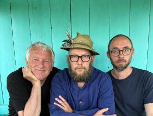 Drei Männer sitzen an einem Holztisch, vor dem eine Konzertzither steht. Der Mann in der Mitte trägt einen Federhut und eine Brille; der Mann links lächelt, und der Mann rechts hat einen Bart und eine Brille. Türkisfarbene Wand im Hintergrund.