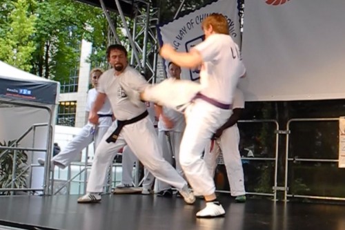 Zwei Kampfsportler in weißen Uniformen und mit schwarzen Gürteln kämpfen auf einer Bühne; einer tritt hoch in Richtung des anderen, der den Tritt blockt. Andere in ähnlichen Uniformen schauen im Hintergrund zu. Hinter ihnen sind Bäume und ein Gebäude zu sehen.