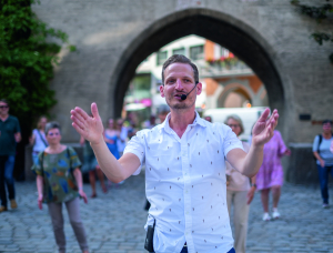 Tanzmeister Magnus Kaindl gestikuliert mit den Händen, während er im Freien vor einem steinernen Torbogen steht. Hinter ihm sieht eine Gruppe von Menschen seinen Anweisungen zu und führt dazu Tanzfiguren auf.