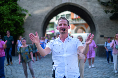 Tanzmeister Magnus Kaindl gestikuliert mit den Händen, während er im Freien vor einem steinernen Torbogen steht. Hinter ihm sieht eine Gruppe von Menschen seinen Anweisungen zu und führt dazu Tanzfiguren auf.