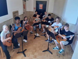 Neun Jugendliche sitzen im Halbkreis in einem Klassenzimmer, jeder hält eine Gitarre in der Hand und schaut in die Kamera. Vor ihnen stehen Notenständer mit Notenblättern. Der Raum hat helle Wände und einen Parkettboden.