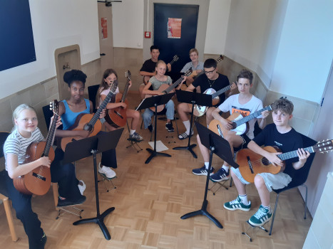 Neun Jugendliche sitzen im Halbkreis in einem Klassenzimmer, jeder hält eine Gitarre in der Hand und schaut in die Kamera. Vor ihnen stehen Notenständer mit Notenblättern. Der Raum hat helle Wände und einen Parkettboden.