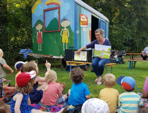 Eine Frau liest einer Gruppe kleiner Kinder ein Bilderbuch vor, die draußen im Gras sitzen, in der Nähe eines bunten Anhängers mit Zeichnungen von Kindern, Bäumen und einem Regenbogen. Die Szene ist hell und sonnig.
