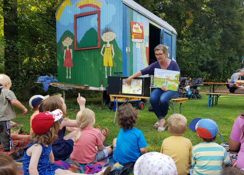 Eine Frau liest einer Gruppe kleiner Kinder ein Bilderbuch vor, die draußen im Gras sitzen, in der Nähe eines bunten Anhängers mit Zeichnungen von Kindern, Bäumen und einem Regenbogen. Die Szene ist hell und sonnig.