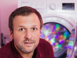 Ein Mann mit kurzen braunen Haaren und Bart steht vor einer offenen Waschmaschine mit bunten Lichtern darin und sieht leicht überrascht oder neugierig aus.