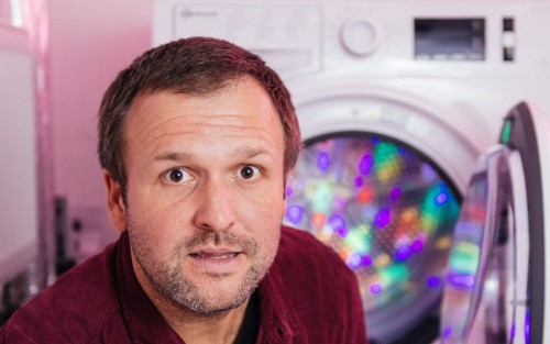 Ein Mann mit kurzen braunen Haaren und Bart steht vor einer offenen Waschmaschine mit bunten Lichtern darin und sieht leicht überrascht oder neugierig aus.