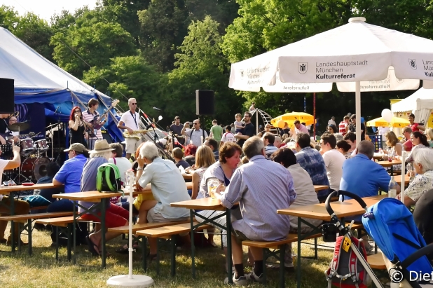 Menschen sitzen an Holztischen unter großen weißen Sonnenschirmen und genießen Essen und Getränke auf einem lebhaften Fest oder Jahrmarkt. Im Hintergrund spielt eine Band unter einem blau-weißen Zelt auf der Bühne. Bäume und Sonnenschein runden die Szene ab.