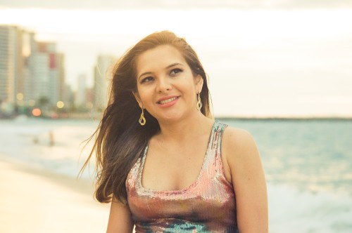 Eine Frau mit langen braunen Haaren, einem glänzenden ärmellosen Top und baumelnden Ohrringen steht an einem Strand. Im Hintergrund sind Meereswellen und Stadtgebäude verschwommen. Sie lächelt und blickt leicht zur Seite.