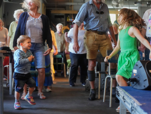 Eine Gruppe von Menschen unterschiedlichen Alters, darunter Kinder und Erwachsene, tanzt und lächelt zusammen in einem Haus. Ein kleiner Junge in einem gestreiften Hemd und ein Mädchen in einem grünen Kleid stehen im Vordergrund und genießen die lebhafte Atmosphäre.