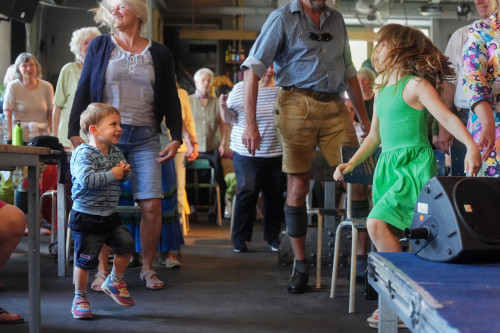 Eine Gruppe von Menschen unterschiedlichen Alters, darunter Kinder und Erwachsene, tanzt und lächelt zusammen in einem Haus. Ein kleiner Junge in einem gestreiften Hemd und ein Mädchen in einem grünen Kleid stehen im Vordergrund und genießen die lebhafte Atmosphäre.