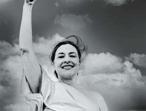 Eine Frau in einem toga-ähnlichen Kleidungsstück steht im Freien, lächelt selbstbewusst mit einem erhobenen Arm und hält einen gestreiften Gegenstand vor einem dramatischen Wolkenhimmel in einem Schwarz-Weiß-Foto.