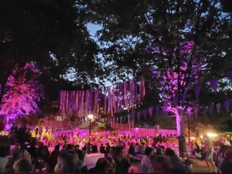 Eine lebhafte Abendveranstaltung im Freien mit Menschen unter Bäumen, die mit lila Lichtern, hängenden Luftschlangen und dreieckigen Wimpeln geschmückt sind. Der Himmel ist dämmrig, und die Umgebung ist von festlicher Atmosphäre erfüllt.