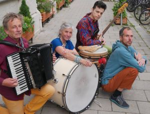 Drei Musiker und eine Musikerin posieren im Freien auf einem Bürgersteig mit ihren Instrumenten: einem Akkordeon, einer großen Trommel, einer Bassgitarre und Bongos. Im Hintergrund säumen Topfpflanzen und Fahrräder die Straße.