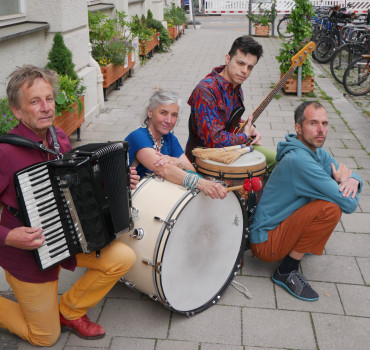 Drei Musiker und eine Musikerin posieren im Freien auf einem Bürgersteig mit ihren Instrumenten: einem Akkordeon, einer großen Trommel, einer Bassgitarre und Bongos. Im Hintergrund säumen Topfpflanzen und Fahrräder die Straße.