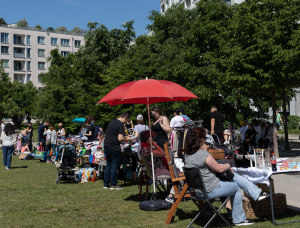 An einem sonnigen Tag stöbern die Menschen auf einem Flohmarkt im Freien und verkaufen ihre Waren. Stände mit Kleidung und Waren säumen das Gras, und ein roter Regenschirm spendet einem Tisch Schatten. Im Hintergrund sind Bäume und moderne Gebäude zu sehen.