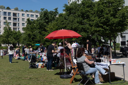 An einem sonnigen Tag stöbern die Menschen auf einem Flohmarkt im Freien und verkaufen ihre Waren. Stände mit Kleidung und Waren säumen das Gras, und ein roter Regenschirm spendet einem Tisch Schatten. Im Hintergrund sind Bäume und moderne Gebäude zu sehen.