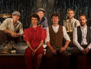 Sechs Personen in Vintage-Kleidung sitzen und knien auf einer Bühne. Musikinstrumente sind zu sehen, hinter ihnen ein funkelnder schwarzer Vorhang. Zwei Männer halten Blechblasinstrumente, in der Mitte steht eine Frau in einem roten Kleid.