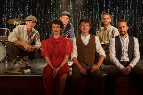 Sechs Personen in Vintage-Kleidung sitzen und knien auf einer Bühne. Musikinstrumente sind zu sehen, hinter ihnen ein funkelnder schwarzer Vorhang. Zwei Männer halten Blechblasinstrumente, in der Mitte steht eine Frau in einem roten Kleid.