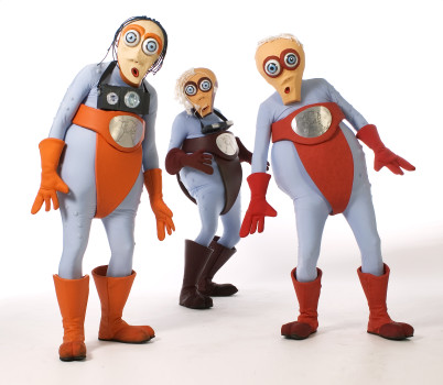 Drei Personen in cartoonhaften Kostümen mit Käferaugen, orangefarbenen Handschuhen, Stiefeln und roten oder braunen Slips posieren vor einem weißen Hintergrund. Alle haben einen übertriebenen Gesichtsausdruck und große, runde Silbermedaillons auf der Brust.