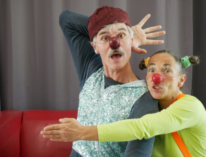 Zwei Erwachsene, die als Clowns mit roten Nasen verkleidet sind, posieren spielerisch. Der Mann trägt ein glitzerndes Oberteil und einen roten Hut, während die Frau in einem hellen Outfit ihn von hinten umarmt. Beide haben einen lustigen Gesichtsausdruck und sind bunt geschminkt.