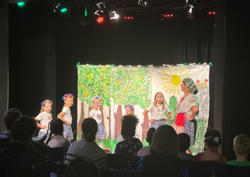 Eine Gruppe kleiner Kinder in geblümten Röcken und Stirnbändern tritt vor einer farbenfrohen Kulisse mit Bäumen und einer Sonne auf der Bühne auf, während ein Publikum in einem schwach beleuchteten Raum zusieht.