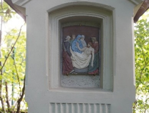 Kleiner Schrein im Freien mit einem Relief, das mehrere Personen um eine sitzende Figur herum darstellt, eingebettet in eine helle Struktur mit einem Kreuz und der römischen Zahl IV über dem Relief, umgeben von grünem Laub.