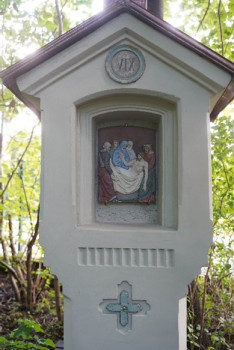 Kleiner Schrein im Freien mit einem Relief, das mehrere Personen um eine sitzende Figur herum darstellt, eingebettet in eine helle Struktur mit einem Kreuz und der römischen Zahl IV über dem Relief, umgeben von grünem Laub.