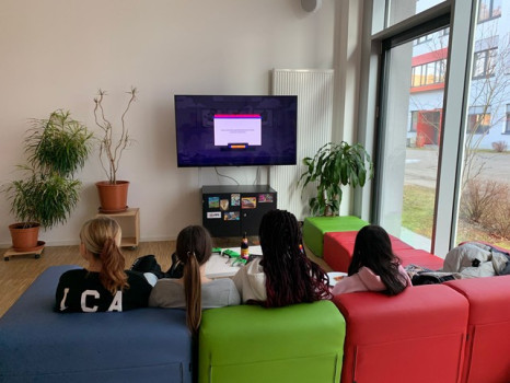 Vier Personen sitzen auf bunten Sofas vor einem großen Fernsehbildschirm in einem hellen Raum mit großen Fenstern, Topfpflanzen und Holzböden. Der Fernseher zeigt eine Nachricht mit einem lila Balken und einer gelben Taste an.