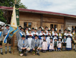 Eine Gruppe von Menschen in traditioneller bayerischer Kleidung posiert vor einem Holzgebäude, Männer, Frauen und Kinder halten Fahnen und tragen Hüte und Schärpen.