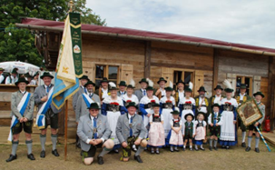 Eine Gruppe von Menschen in traditioneller bayerischer Kleidung posiert vor einem Holzgebäude, Männer, Frauen und Kinder halten Fahnen und tragen Hüte und Schärpen.