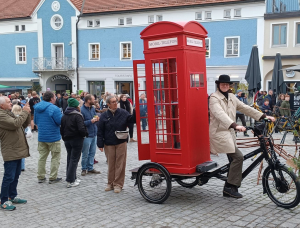Eine Person im Anzug und mit Bowlerhut fährt auf einem Dreirad, an dem eine große rote Telefonzelle im britischen Stil befestigt ist, während eine Menschenmenge auf einem Stadtplatz zusieht und Fotos macht.