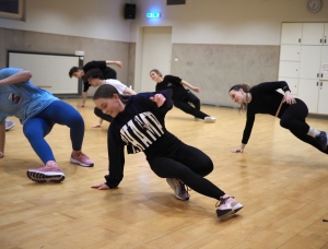 Eine Gruppe Tänzer in legerer Sportkleidung übt Bodenbewegungen in einem hell erleuchteten Tanzstudio mit Holzboden und weißen Wänden. Sie befinden sich mitten in der Bewegung, stützen sich ab und balancieren mit einem Arm auf dem Boden.