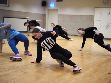 Eine Gruppe Tänzer in legerer Sportkleidung übt Bodenbewegungen in einem hell erleuchteten Tanzstudio mit Holzboden und weißen Wänden. Sie befinden sich mitten in der Bewegung, stützen sich ab und balancieren mit einem Arm auf dem Boden.