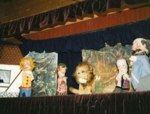 Eine Puppentheaterbühne mit fünf Puppen, darunter ein Löwe, zwei menschliche Figuren und zwei kleinere Figuren, vor einer gemalten Waldkulisse mit dunklen Vorhängen, die die Szene einrahmen.