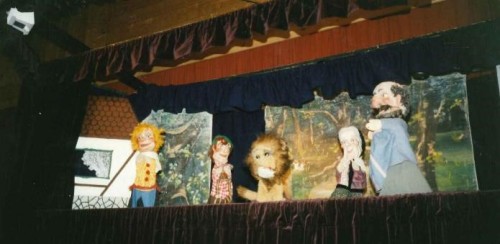 Eine Puppentheaterbühne mit fünf Puppen, darunter ein Löwe, zwei menschliche Figuren und zwei kleinere Figuren, vor einer gemalten Waldkulisse mit dunklen Vorhängen, die die Szene einrahmen.