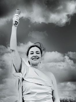 Eine Frau in einem toga-ähnlichen Kleidungsstück steht im Freien, lächelt selbstbewusst mit einem erhobenen Arm und hält einen gestreiften Gegenstand vor einem dramatischen Wolkenhimmel in einem Schwarz-Weiß-Foto.