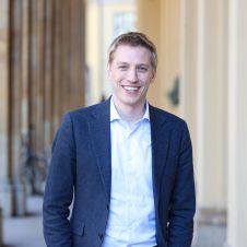 Dominik Krause steht in einem marineblauen Blazer und einem weißen Hemd lächelnd in den Hofgarten-Akaden
