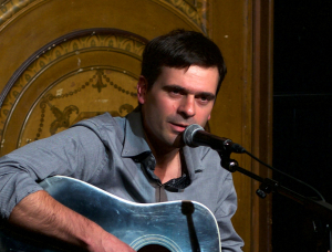 Ein Mann sitzt und spielt eine Akustikgitarre, während er in ein Mikrofon singt. Er trägt ein graues Hemd und eine dunkle Hose und hat einen dekorativen Holzhintergrund im Rücken.