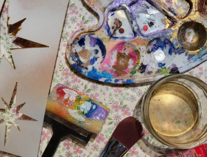 Eine Farbpalette mit bunter Farbe, ein Glas Wasser, ein Pinsel, ein Pinsel, eine Tube rote Acrylfarbe und eine Sternschablone liegen auf einer blumengemusterten Fläche.