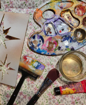 Eine Farbpalette mit bunter Farbe, ein Glas Wasser, ein Pinsel, ein Pinsel, eine Tube rote Acrylfarbe und eine Sternschablone liegen auf einer blumengemusterten Fläche.