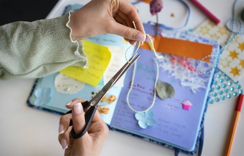 Eine Person schneidet mit einer Schere eine Schnur über einem offenen Sammelalbum durch, das mit verstreuten Papierblumen, Notizen und verschiedenen Bastelmaterialien dekoriert ist.