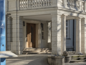 Steinsäulen stützen einen überdachten Eingang mit hölzernen Doppeltüren an einem hellen Steingebäude. Ein blaues Schild mit der Nummer 4 und eine Informationstafel stehen in der Nähe. Schatten und Sonnenlicht schaffen eine ruhige Atmosphäre.
