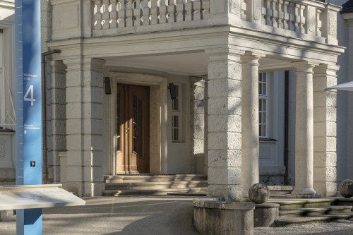 Steinsäulen stützen einen überdachten Eingang mit hölzernen Doppeltüren an einem hellen Steingebäude. Ein blaues Schild mit der Nummer 4 und eine Informationstafel stehen in der Nähe. Schatten und Sonnenlicht schaffen eine ruhige Atmosphäre.